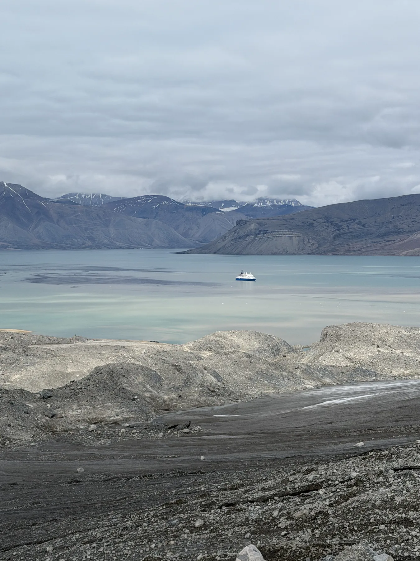 Moraine rubble in foreground, glacial-milk fjord beyond, a ship at anchor for scale