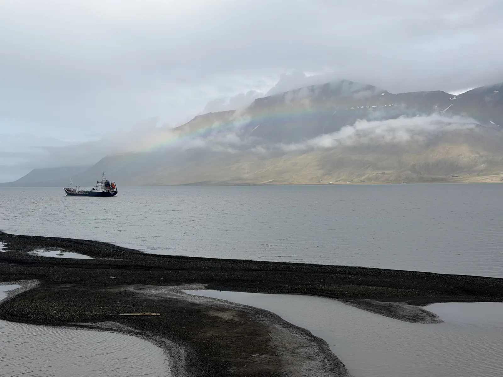 A black sand tidal spit at Longyearbyen with a rainbow over the fjord and a ship